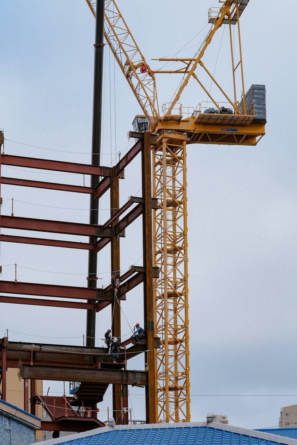 Construction workers stand on a steel building frame near a crane.
